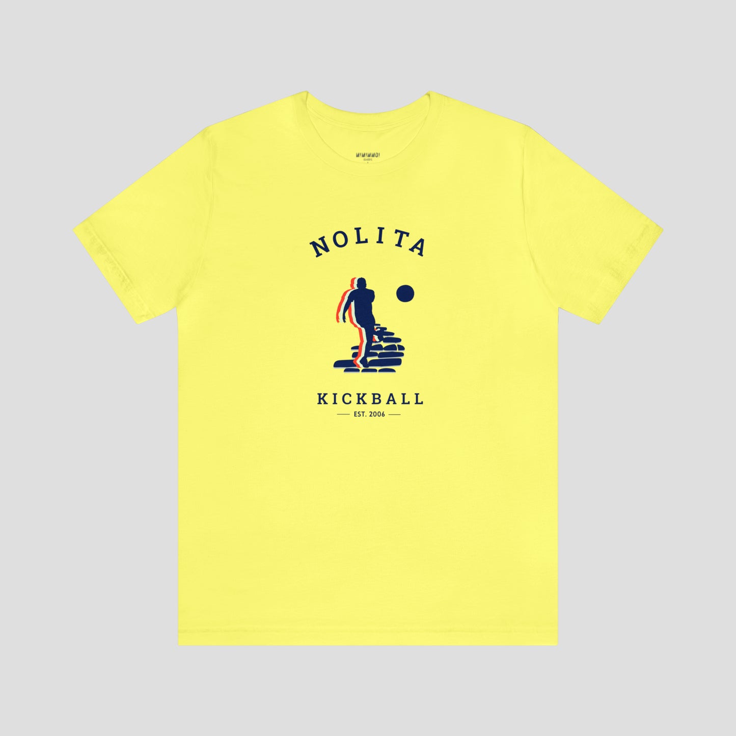 Yellow t-shirt with a colorfully shadowed navy silhouette of person kicking a ball on cobblestone street. The words Nolita Kickball est. 2007 are written in navy athletic font. The word Nolita is curved above the image and the other words are below the image. The full t-shirt image is against a gray background.