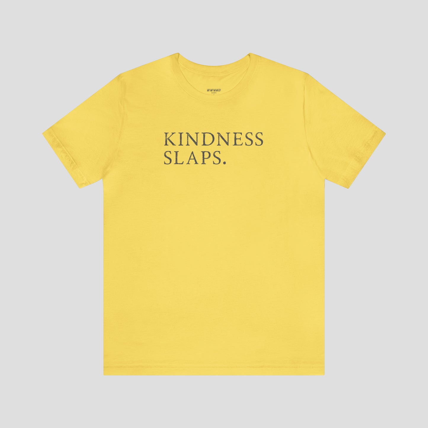 Yellow t-shirt with the words kindness slaps in capitalized gray typeface letters across the chest, against a gray background.