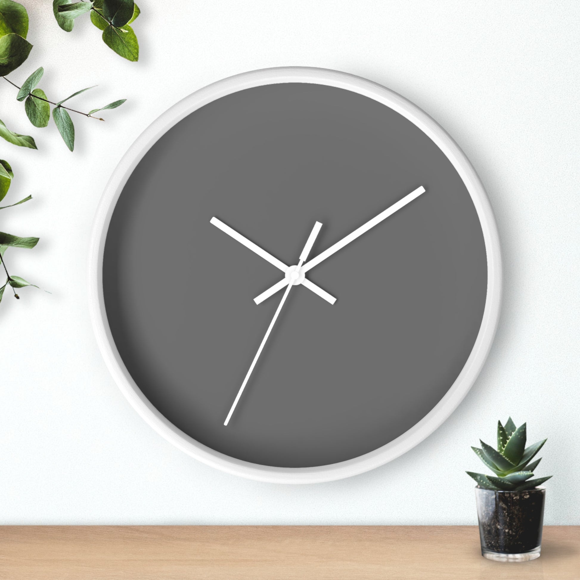 White circular wooden framed clock with dark gray background and white hands with leaves on upper left corner and small cactus on bottom right against a white background.