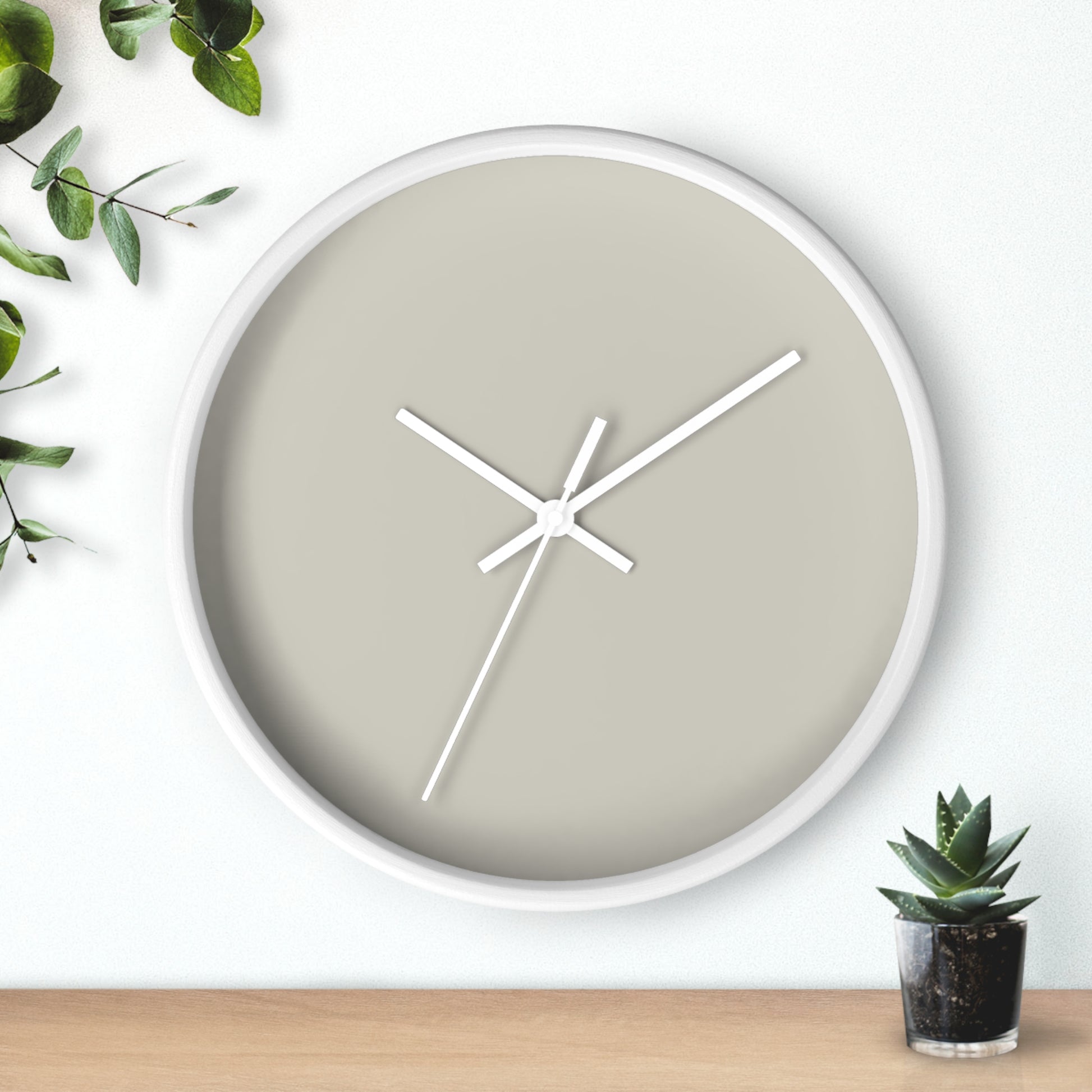 White circular wooden framed clock with tan background and white hands with leaves on upper left corner and small cactus on bottom right against white background.