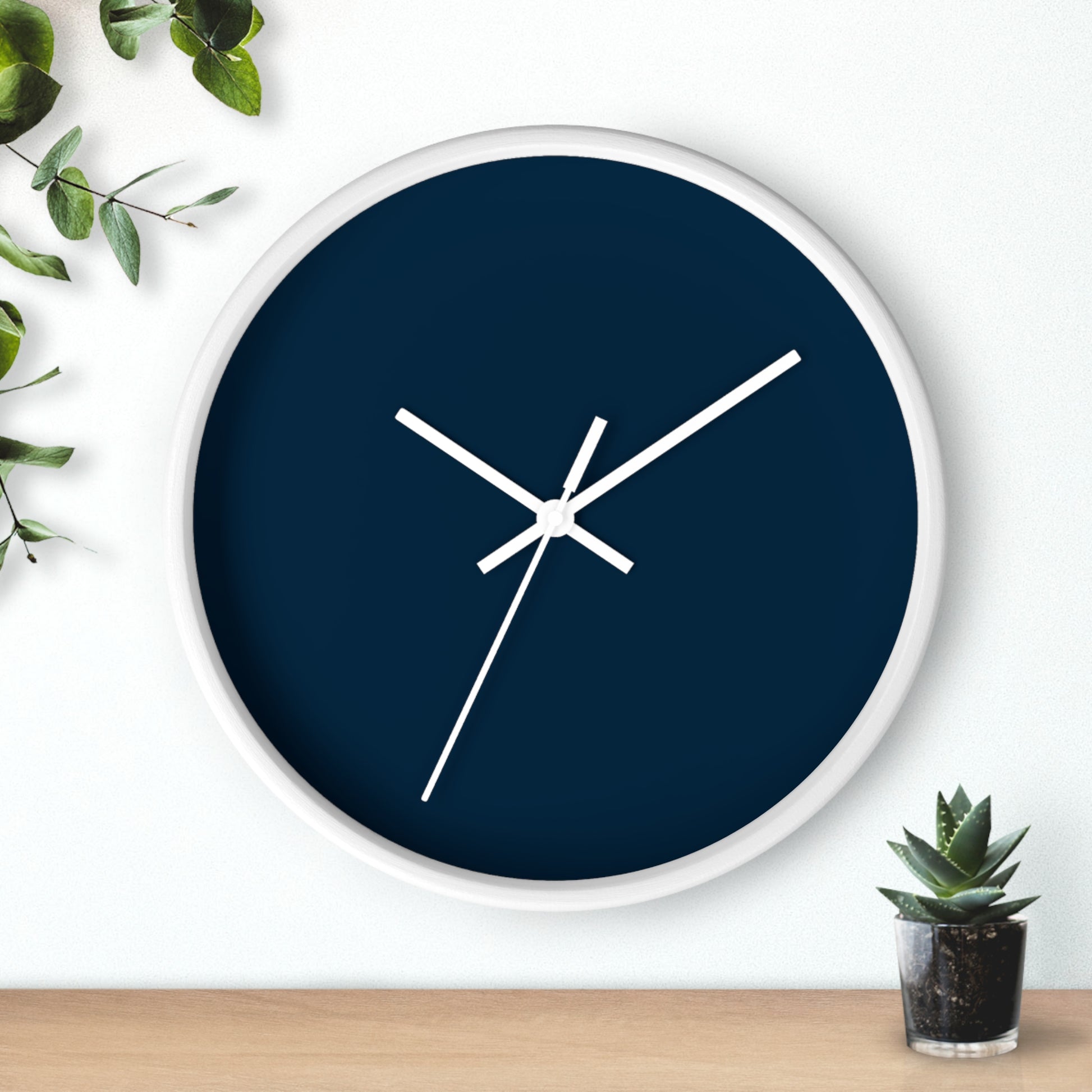 White circular wooden framed clock with navy blue background and white hands with leaves on upper left corner and small cactus on bottom right against a white background.