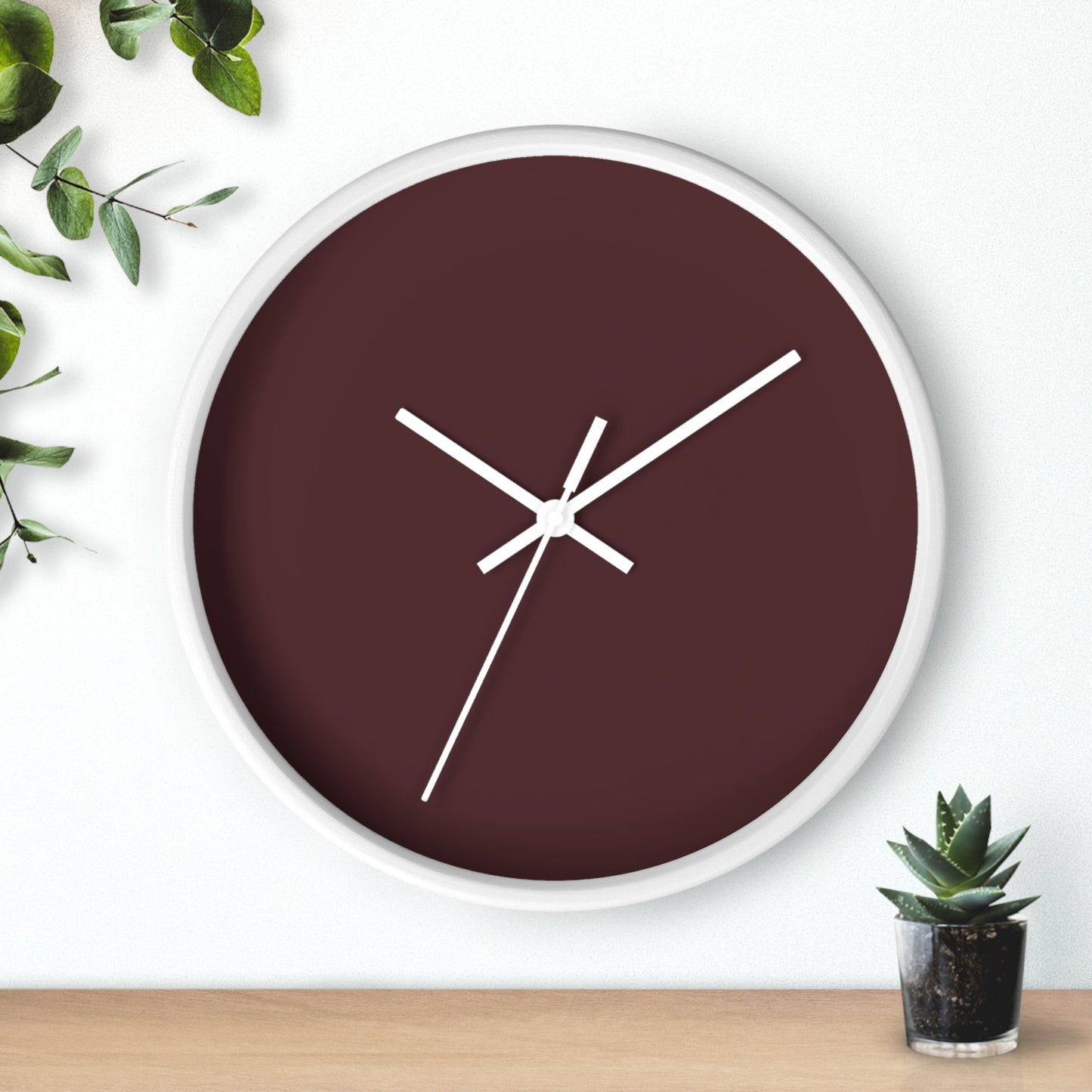 White circular wooden framed clock with maroon colored background and white hands with leaves on upper left corner and small cactus on bottom right against a white background.