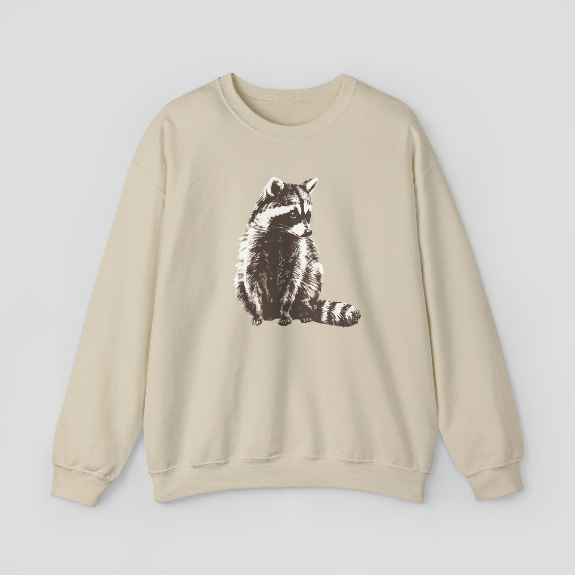 Tan colored sweatshirt with large brown and white illustrated image of a racoon across the chest, against a gray background.