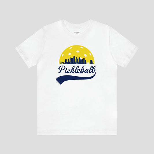 White t-shirt with large image in center of a pickleball behind silhouette of Los Angeles with the word Pickleball written in blue cursive lettering, against a gray background.