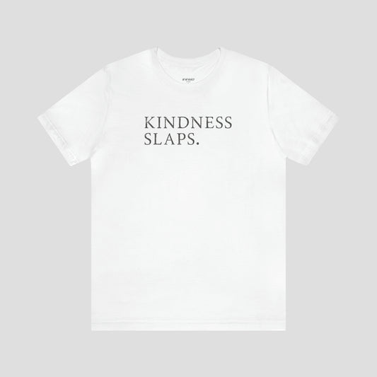 White t-shirt with the words kindness slaps in capitalized gray typeface letters across the chest, against a gray background.