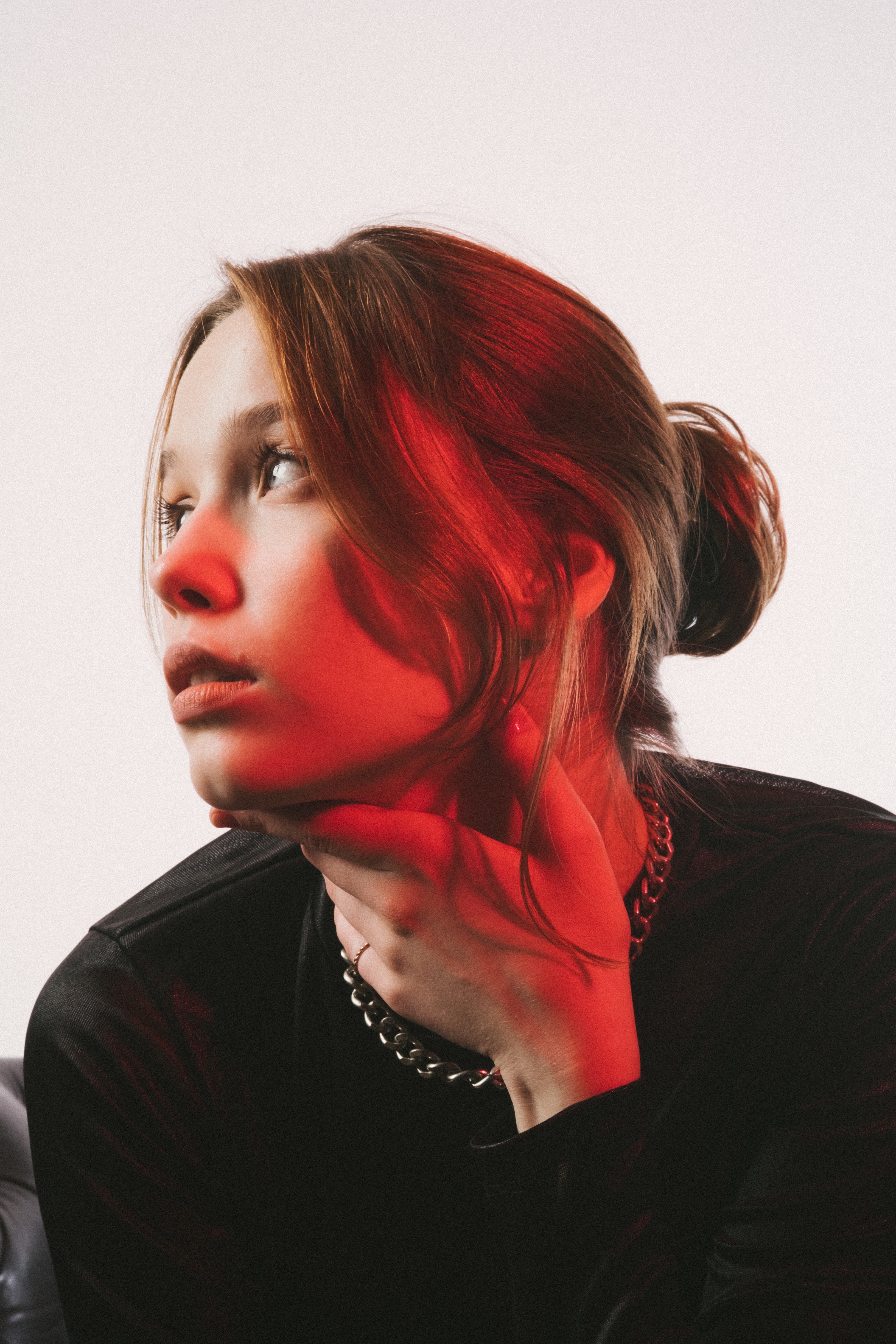 Young woman with hand placed on neck looking to the side. She is wearing a chain and small ponytail.